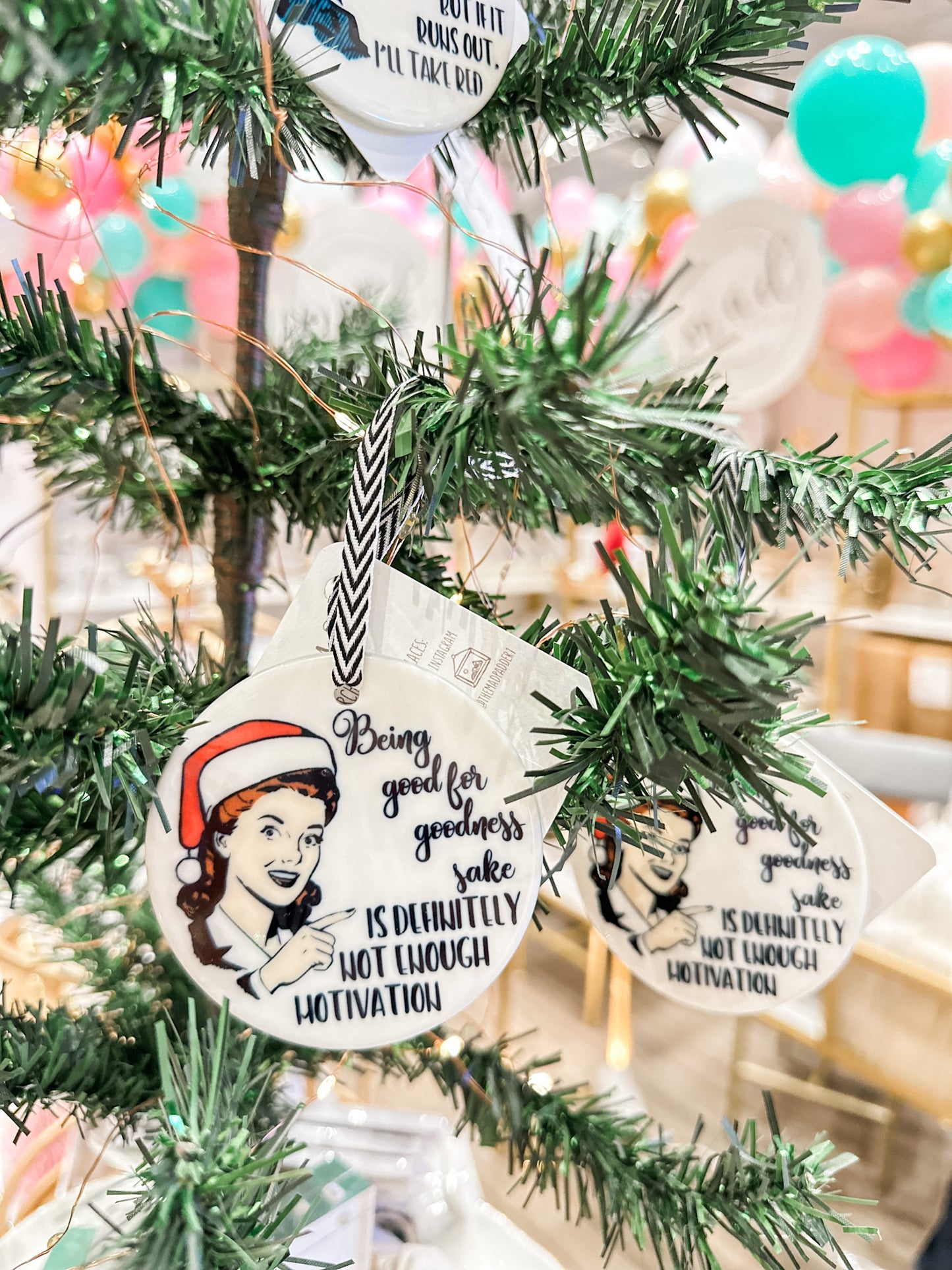 The Mad Padder Witty Women Holiday Ornament  saying "Being good for goodness sake is definitely not enough motivation" featuring a smiling vintage woman and a Santa hat on her head on a round, ceramic ornament within a tree.
