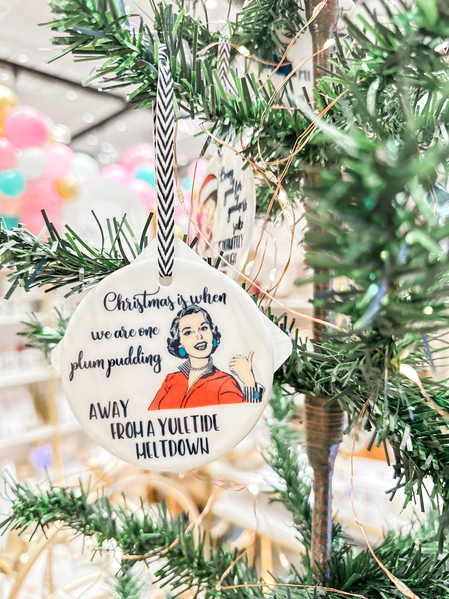 The Mad Padder Witty Women Holiday Ornaments "Christmas is when we are one plum pudding away from a yuletide meltdown" on an ornament featuring a smiling vintage woman wearing a red blouse and green earrings on a white, ceramic ornament within a tree.