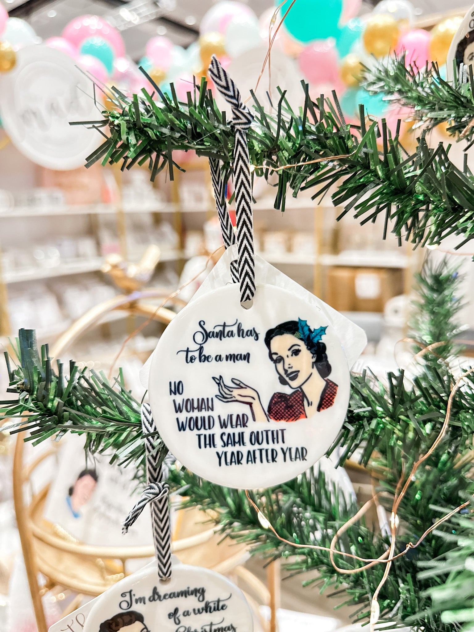 The Mad Padder Witty Women Holiday Ornaments  saying "Santa has to be a man...No woman would wear the same outfit year after year" on an ornament featuring a smiling vintage woman with holly in her hair on a white, round, ceramic ornament within a tree.